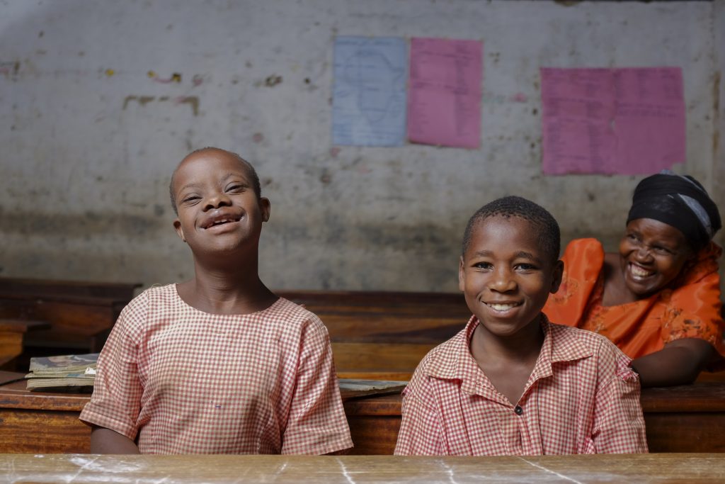 Two children are photographed in a classroom, a woman sits behind them smiling.